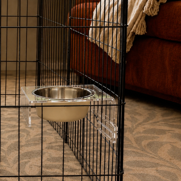 A clear acrylic pet feeder mount holding a stainless steel bowl and beige ceramic feeder makes up the Nook No Tip Bowl station,  attached to a black metal playpen in a living room.
